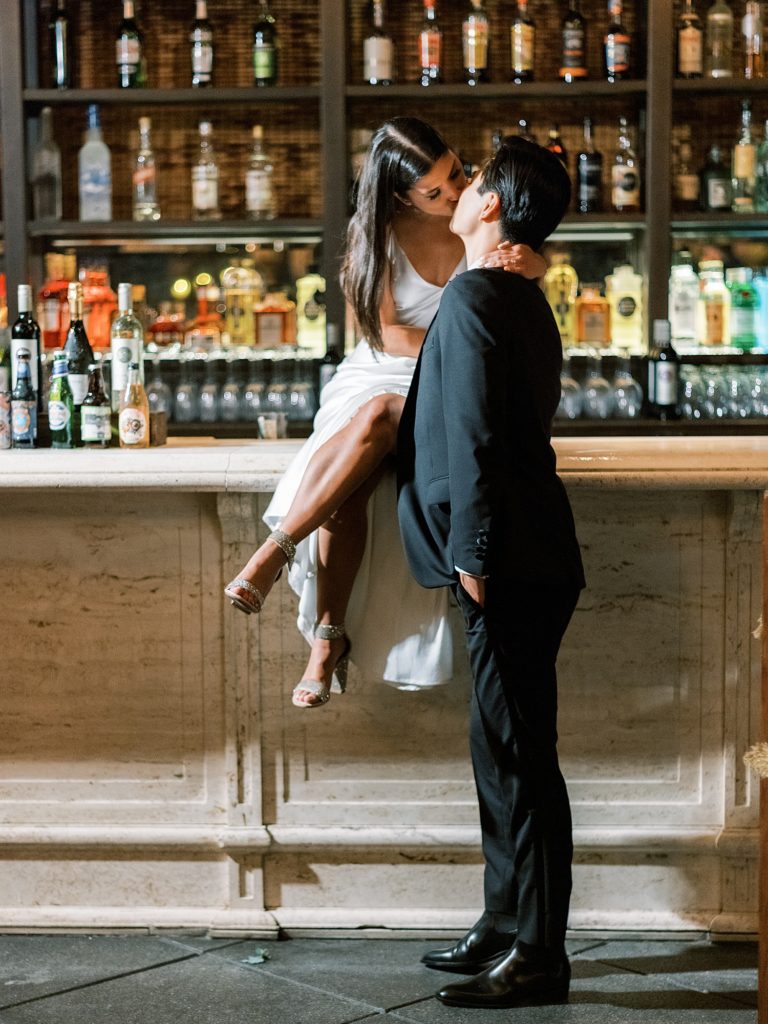Bride in silk dress and groom in tux kiss on bar.