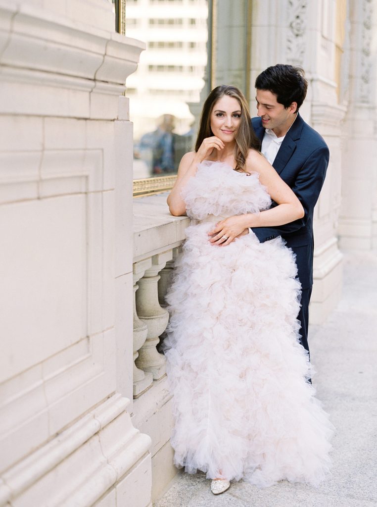 Man in suit and woman in beautiful designer gown leaning against a stone wall.