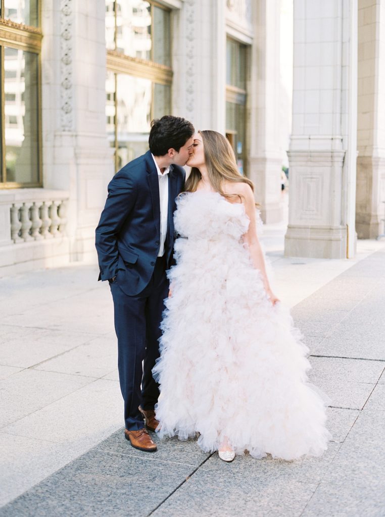 Woman in tulle gown and man in suit kiss in front of old stone building in Chicago.