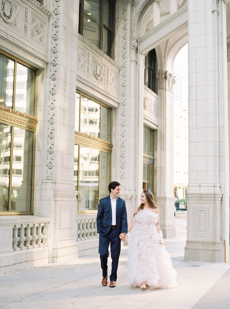 Man in suit and woman in a tulle gown walk through stone building walkway.
