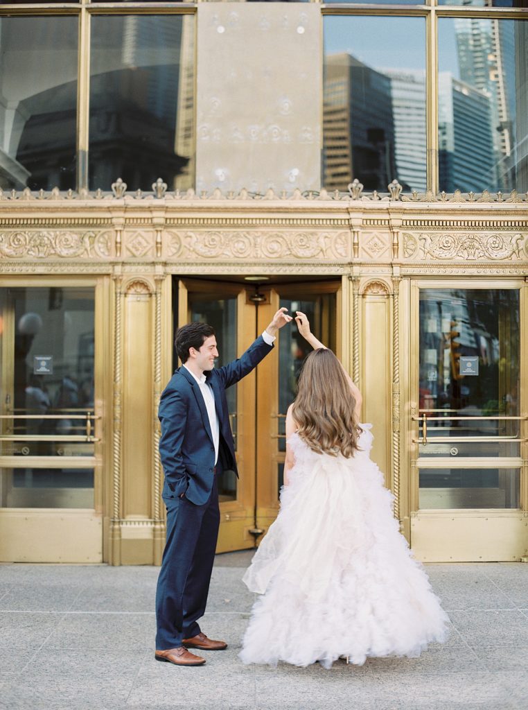 Man twirling woman around in a designer gown in front of ornate gold doors on a Chicago building.