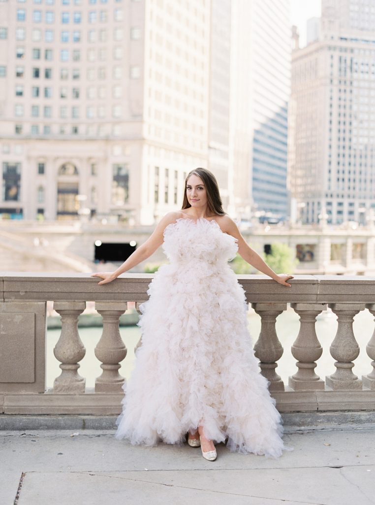 Woman in designer tulle gown, leans agains a stone walk way with city buildings in the background.