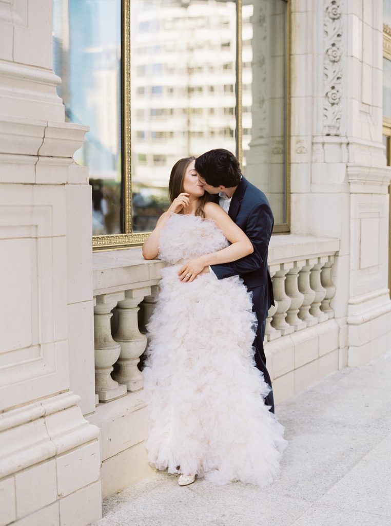Woman in tulle dress and man in suit curl up and kiss next to stone walkway and large windows.