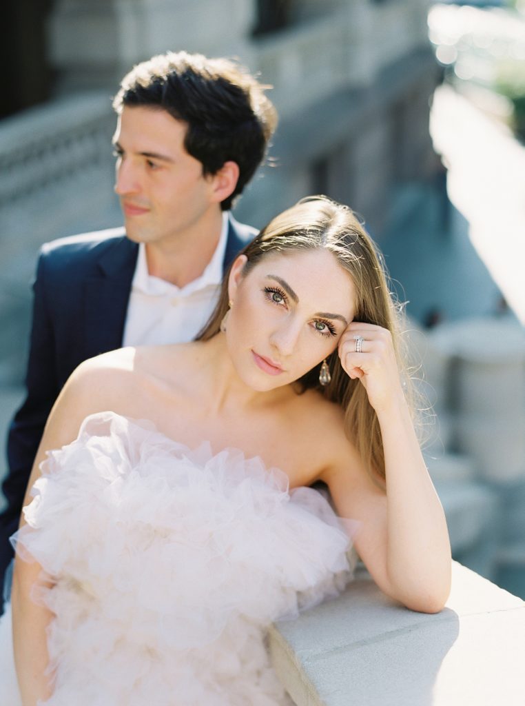 Woman in tulle designer dress leaning on a stone stair railing, looking up at the camera. Her man looking off into the distance behind her.