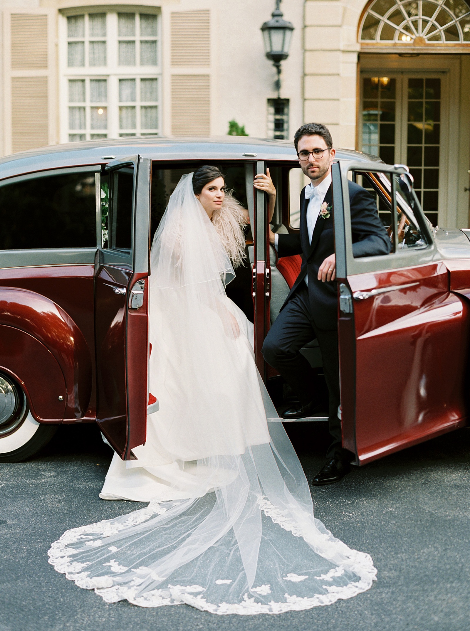 Bride and groom posed around a vintage red car, looking at the camera.