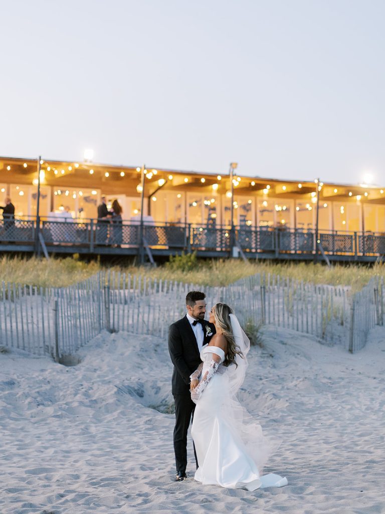 Bride and groom on the beach with the lights from their wedding reception behind them. 