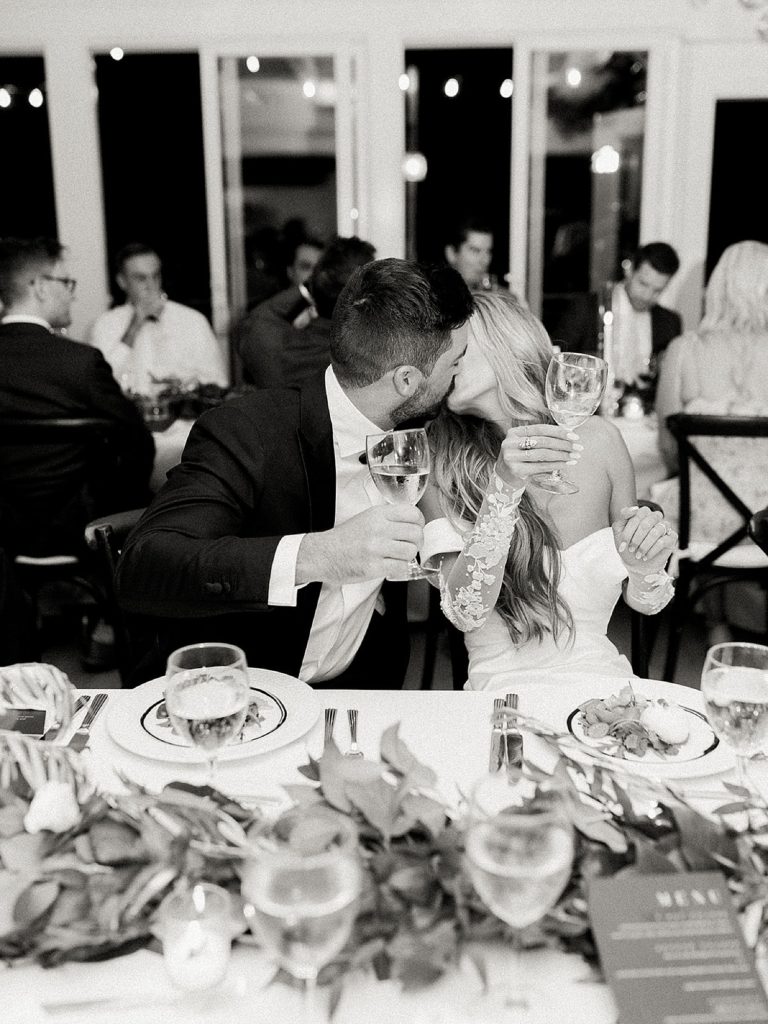Bride and groom kiss during the toasts at their wedding surrounded by their friends and family. 