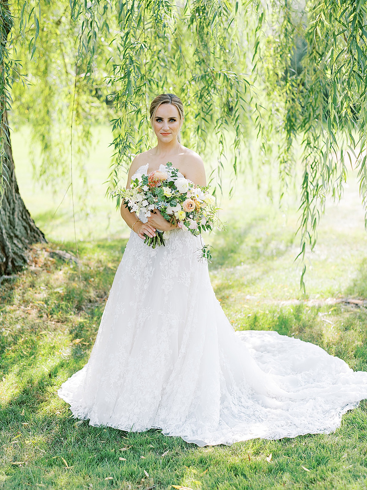 Bride poses in embroidered lace wedding gown holding pastel bouquet in front of weeping willow shot by Sophie Kaye photography